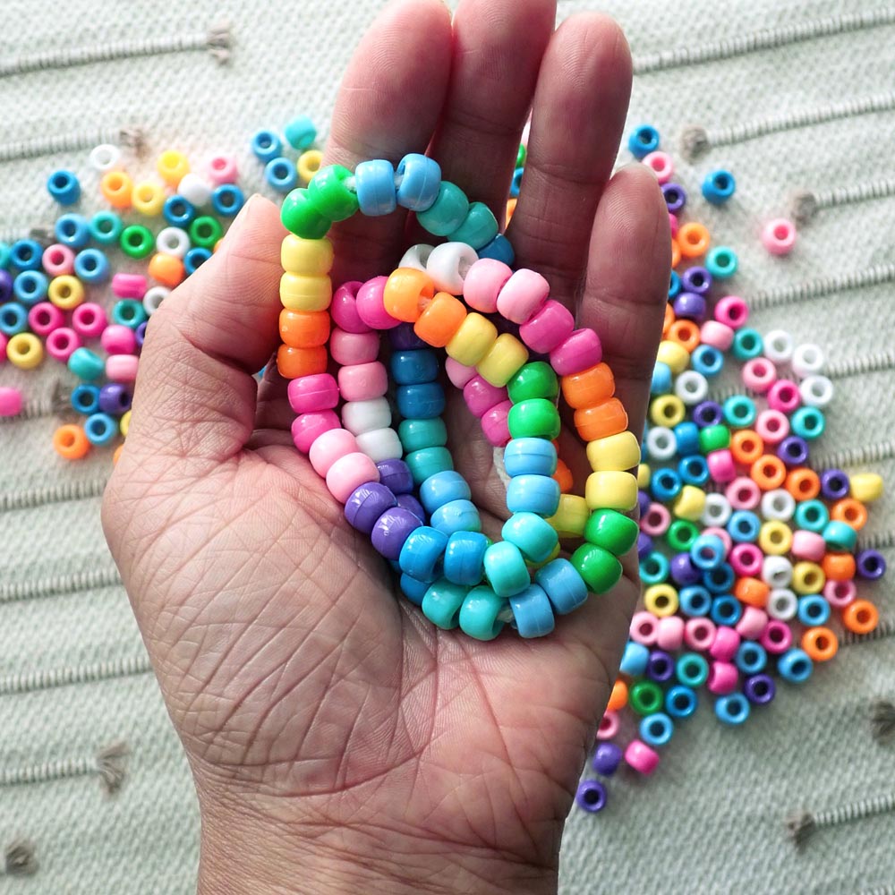 Hand holding a coiled necklace made of colorful pony beads with more beads scattered on a textured surface.