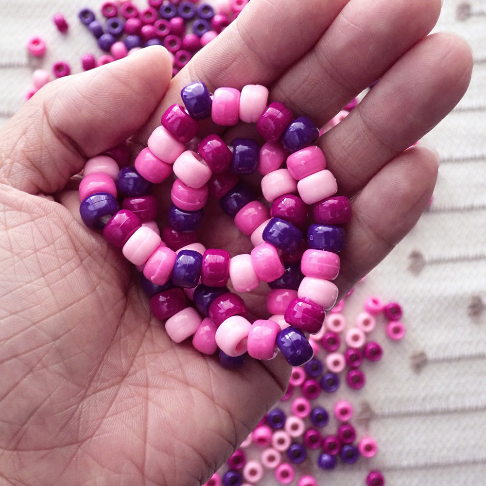 Hand holding a coiled necklace made of pink and purple pony beads with more beads scattered on a textured surface.
