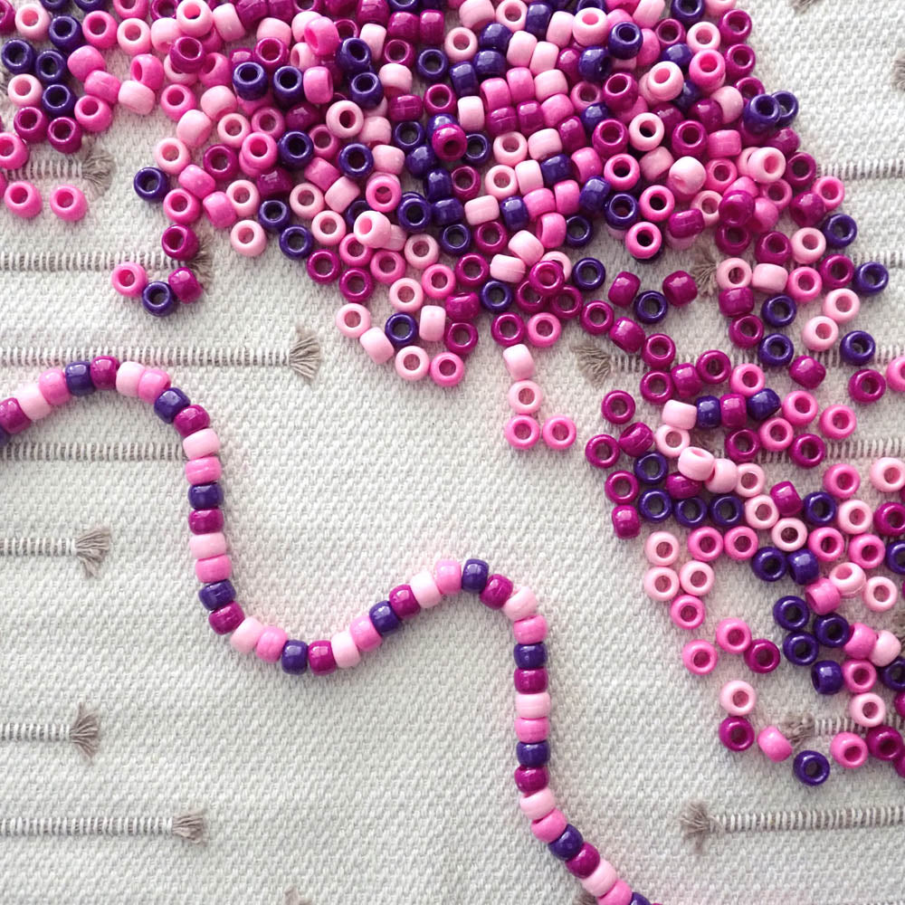 pink & purple color mix of pony beads shown as loose beads and a string of beads on a textured fabric background
